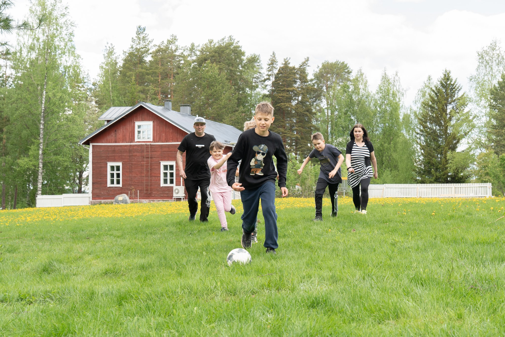 Mies, nainen, kaksi poikaa ja yksi tyttö pelaavat jalkapalloa ulkona nurmikolla. Taustalla näkyy puita ja punainen puutalo.