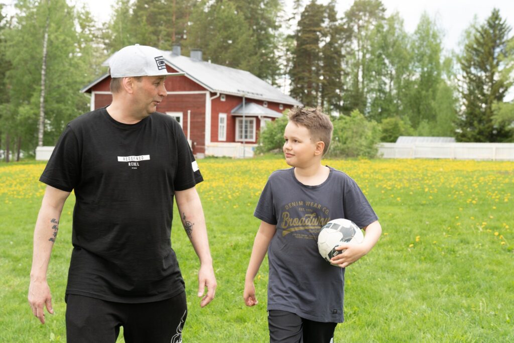 “Aikuinen ja lapsi kävelevät nurmikolla jutellen, lapsella on jalkapallo kainalossa. Taustalla näkyy punainen talo ja kesäinen maisema.