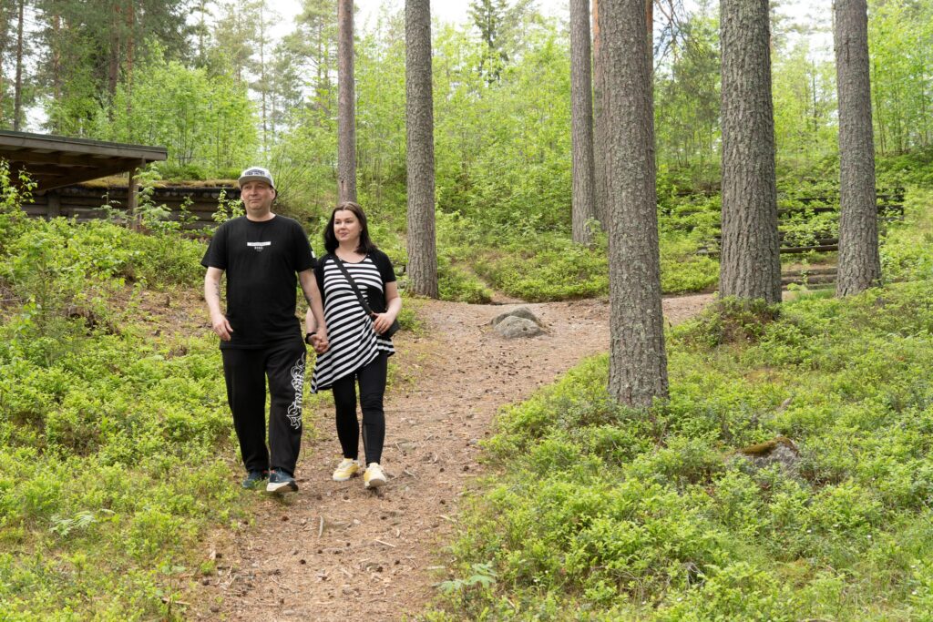 Aikuinen mies ja nainen kävelevät yhdessä metsäpolulla. Heidän ympärillään kohoaa mäntymetsä ja vihreä aluskasvillisuus, taustalla näkyy pieni laavu.