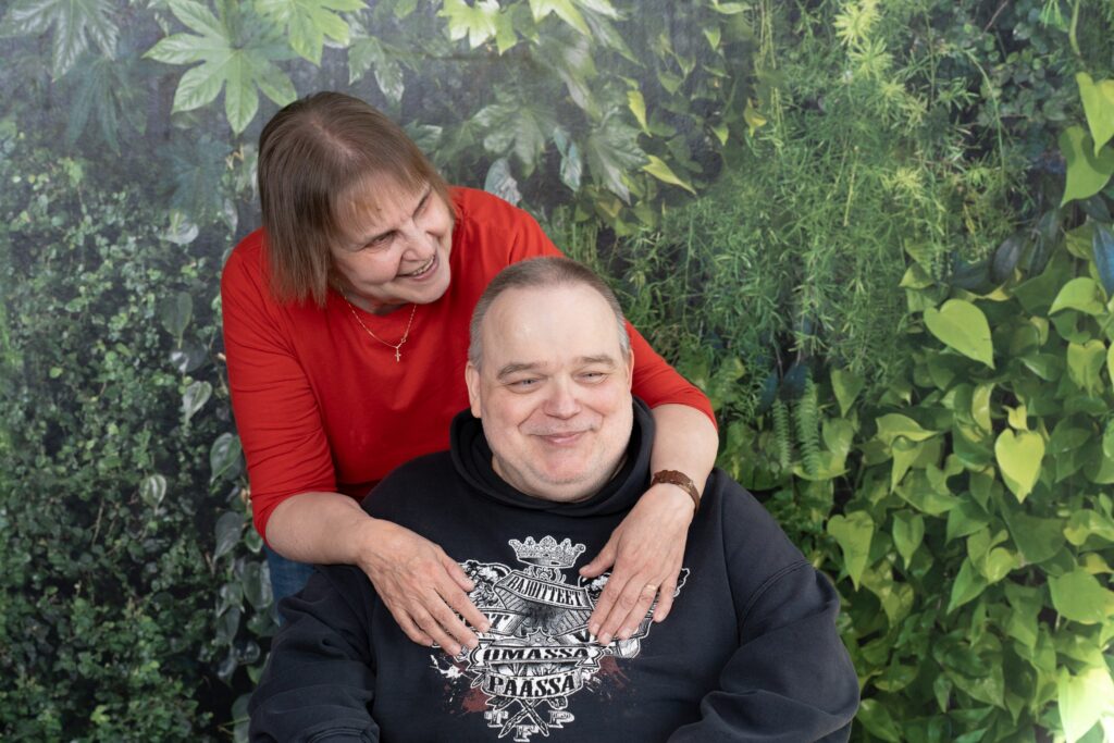 Tummaan pukeutunut mies istuu pyörätuolissa. Punaiseen paitaan pukeutunut nainen seisoo hänen takanaan ja kumartuu halaamaan häntä. Taustalla on vihreä kasvitapetti.