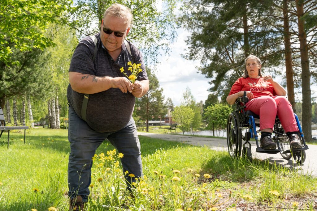 Mies kerää keltaisia kukkia kesäisessä maisemassa, ja taustalla nainen istuu pyörätuolissa hymyillen. Ympärillä vihreää nurmikkoa, puita ja aurinkoinen sää.