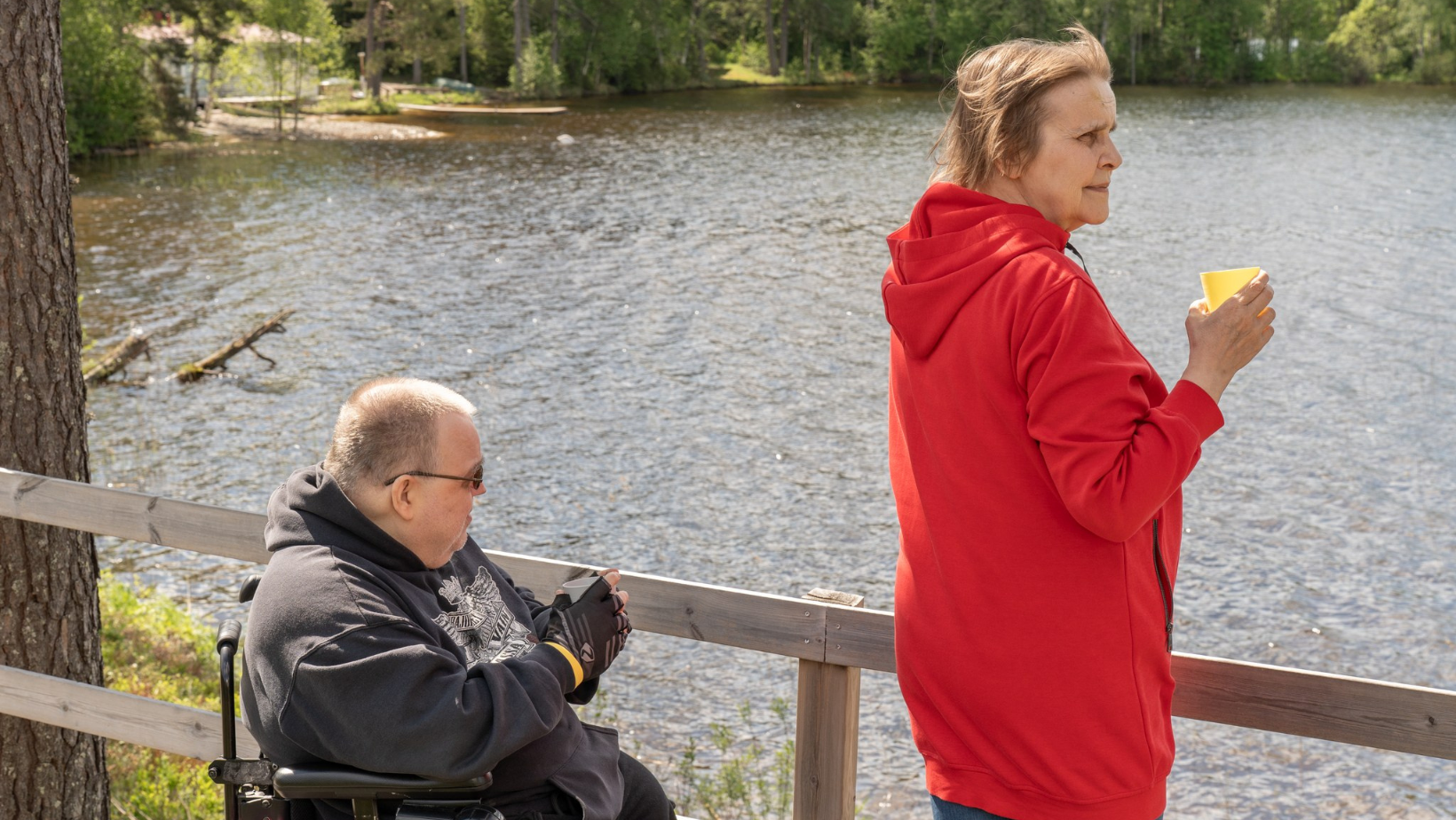 Kaksi aikuista istuu ja seisoo järven rannalla puisen kaiteen äärellä; toinen henkilö käyttää pyörätuolia ja toinen nojaa kaiteeseen juoma kädessä. Aurinkoinen kesäpäivä ja vihreä metsä näkyvät taustalla.